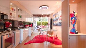 Una cocina moderna con electrodomésticos de acero inoxidable, un comedor con mesa de vidrio y sillas blancas, y una alfombra roja.