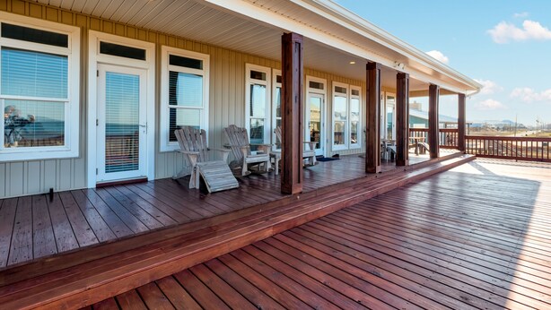 Une terrasse en bois couverte, dotée de plusieurs portes et fenêtres, d’un espace salon extérieur et offrant une vue sur l’océan.