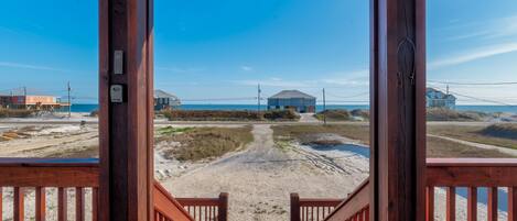 Una terraza de madera que conduce a una playa de arena, con vista despejada al océano y una casa azul distante.