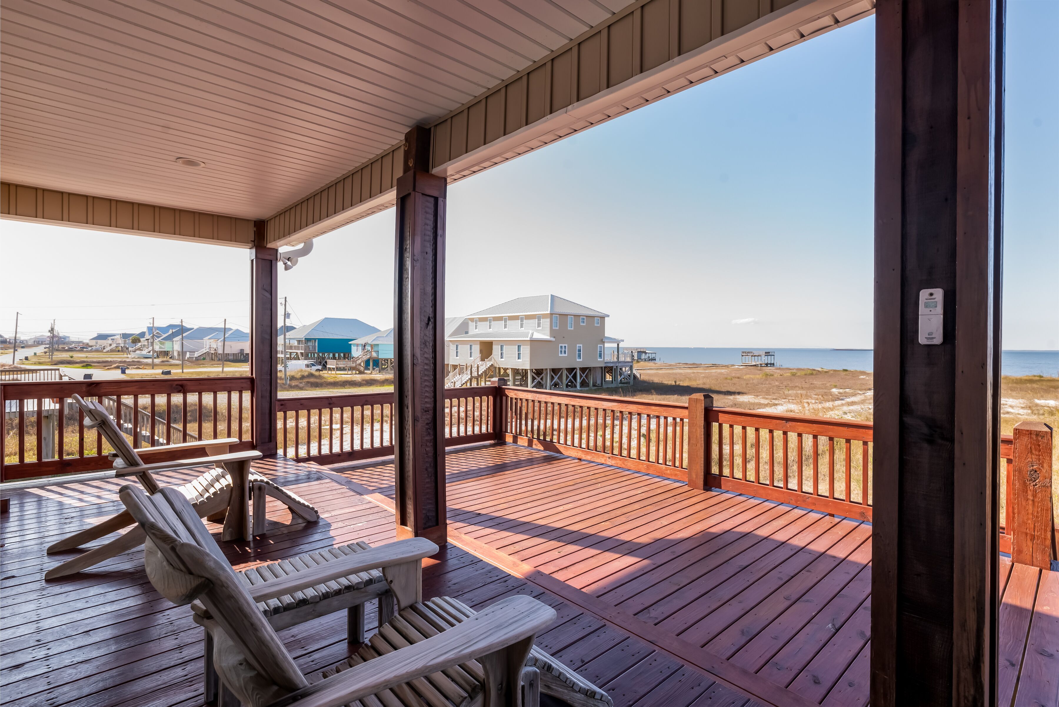A wooden deck with two Adirondack chairs and a table, overlooking a beach and a large building.