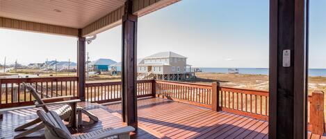 A wooden deck with two Adirondack chairs and a table, overlooking a beach and a large building.