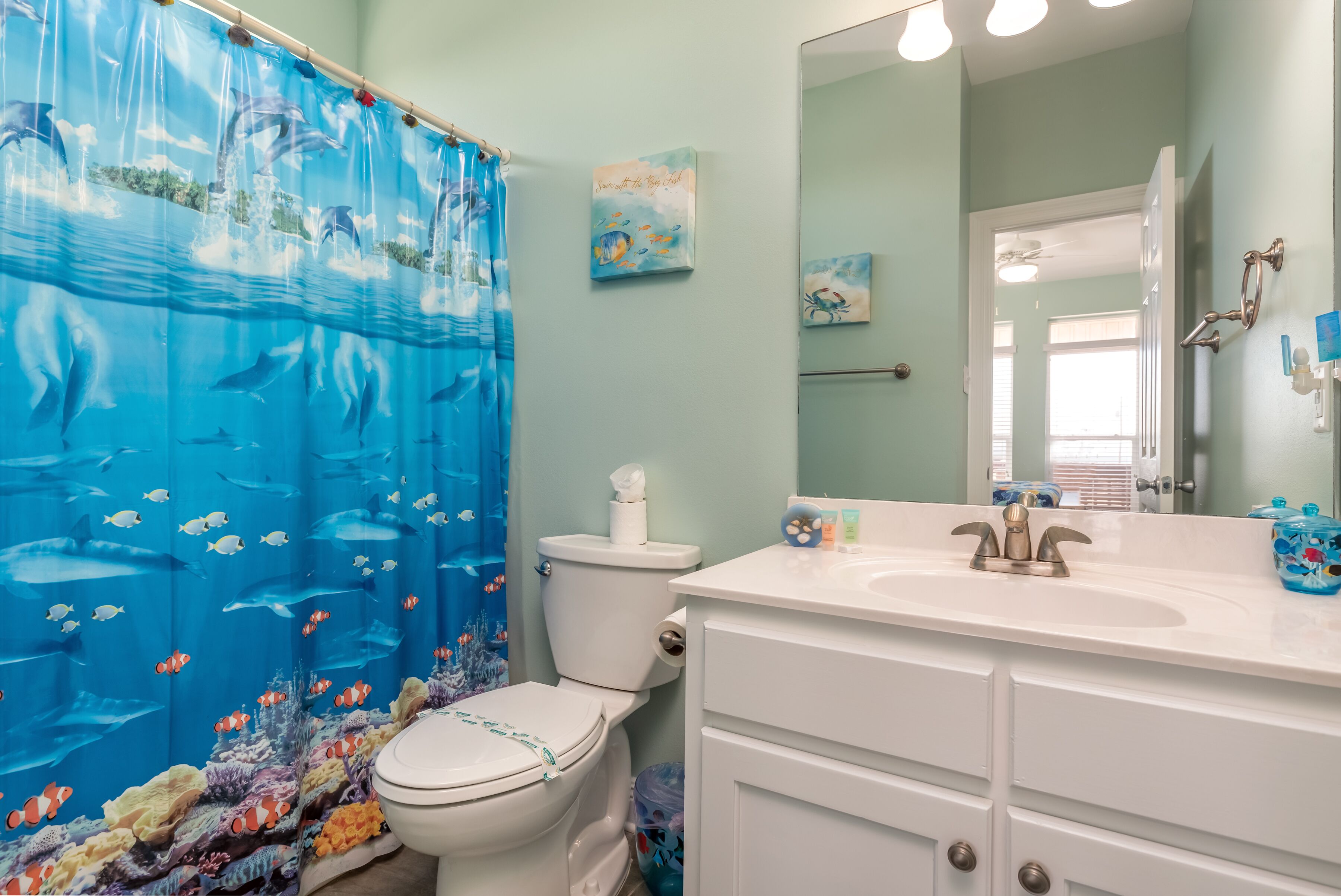 Une salle de bain avec un rideau de douche à motifs de dauphins, des toilettes, un lavabo avec un miroir et un meuble de rangement.