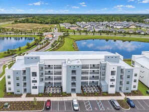 Un immeuble à plusieurs étages, avec un parking à l’avant, entouré d’un lac et d’un quartier résidentiel.