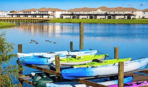 Une rangée de kayaks aux couleurs vives est amarrée à un quai, avec un lac paisible et des maisons en arrière-plan.