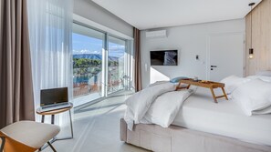 A modern hotel room with a large bed, a laptop on a side table, a TV mounted on the wall, and a balcony with a scenic view.