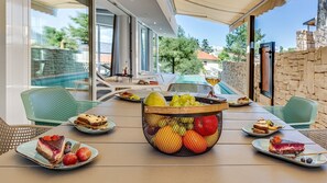 An outdoor dining area with a table set for four, surrounded by wicker chairs. A fruit basket filled with various fruits is placed in the center.