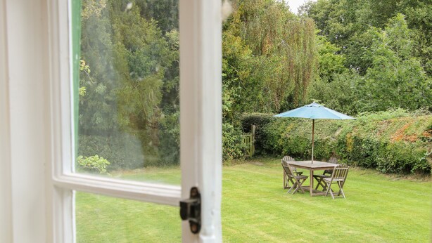 A view through a window of a garden with a wooden table and chairs under a blue umbrella, surrounded by lush greenery. - Denbigh Hall (Leominster)