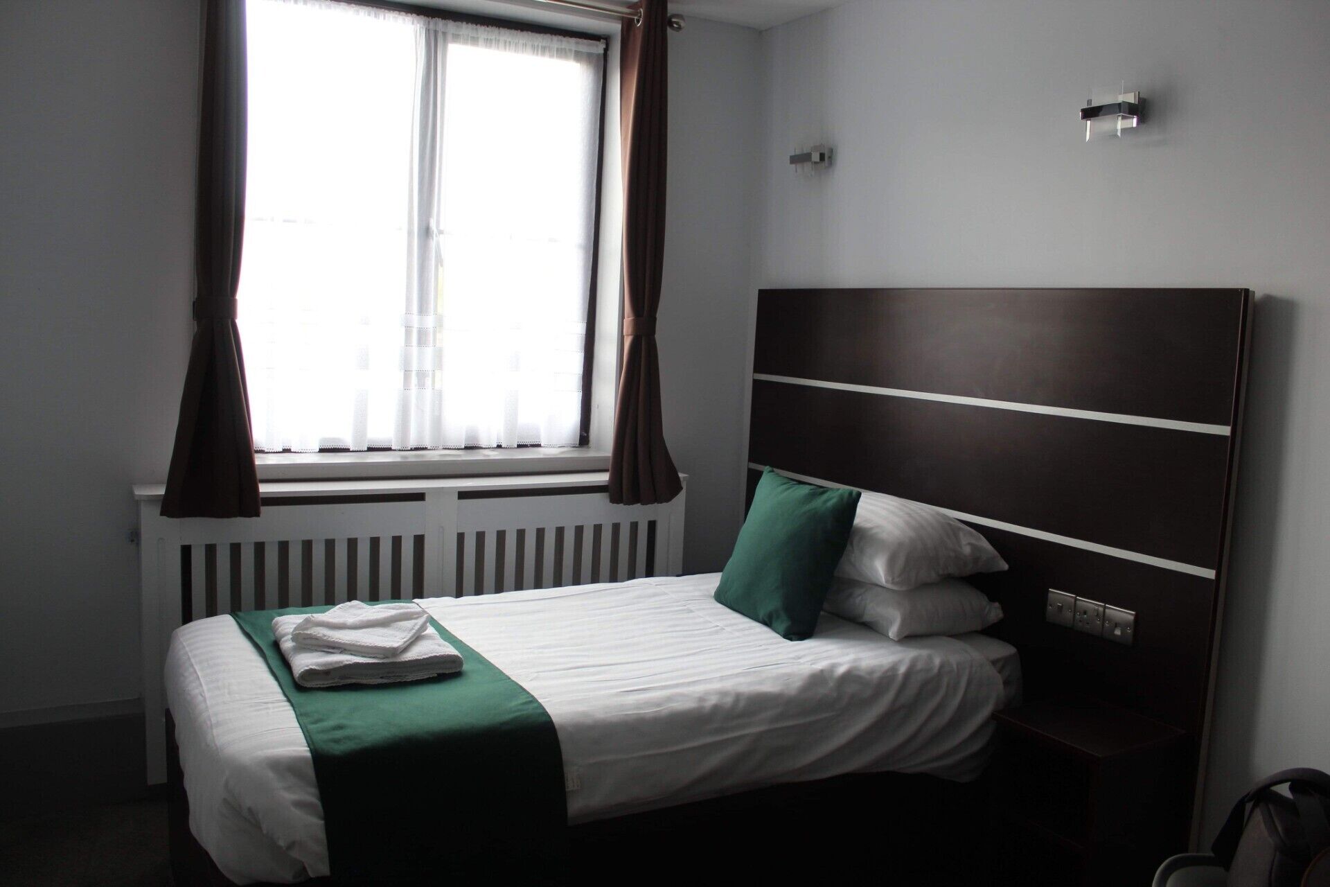 Et hotelværelse med seng, et vindue med gardiner, en radiator og et sengebord.