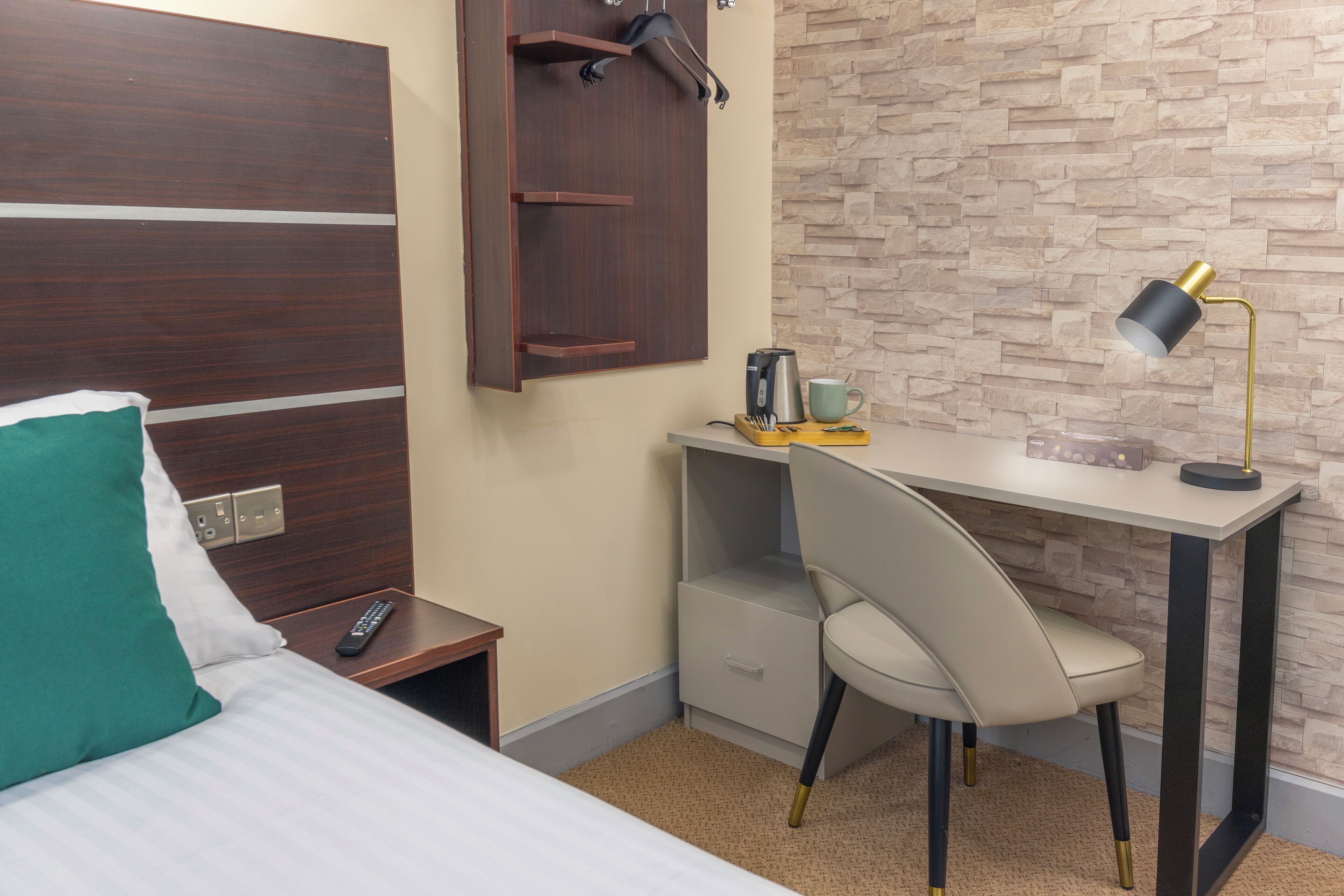 A hotel room with a bed, a desk with a chair, a desk lamp, a coffee maker, and a wall-mounted coat rack.