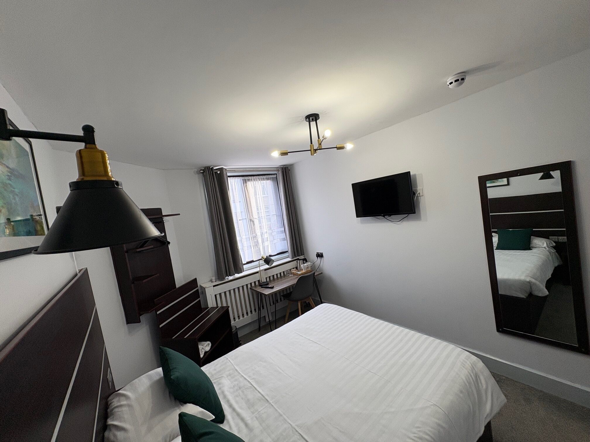 Ein Hotelzimmer mit Bett, Schreibtisch, Stuhl, Lampe, Fernseher, Spiegel und einem fenster mit Vorhängen.