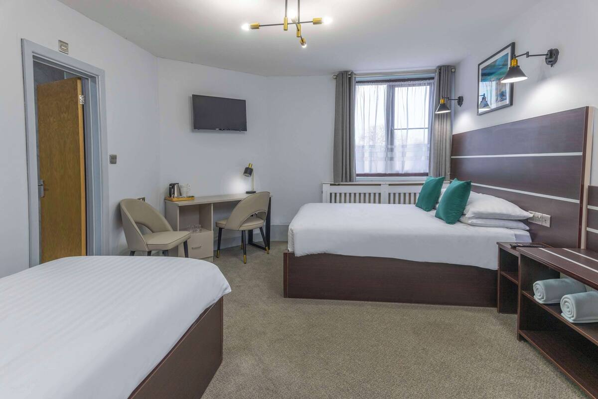 A hotel room with two single beds, a desk with a chair, a TV mounted on the wall, a window with curtains, and a bedside table with towels.