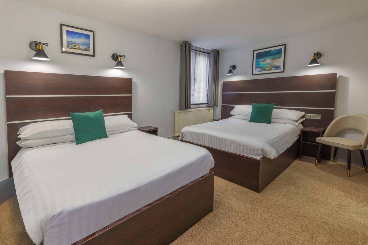 A hotel room with two beds, a chair, a window with curtains, and wall-mounted lamps.