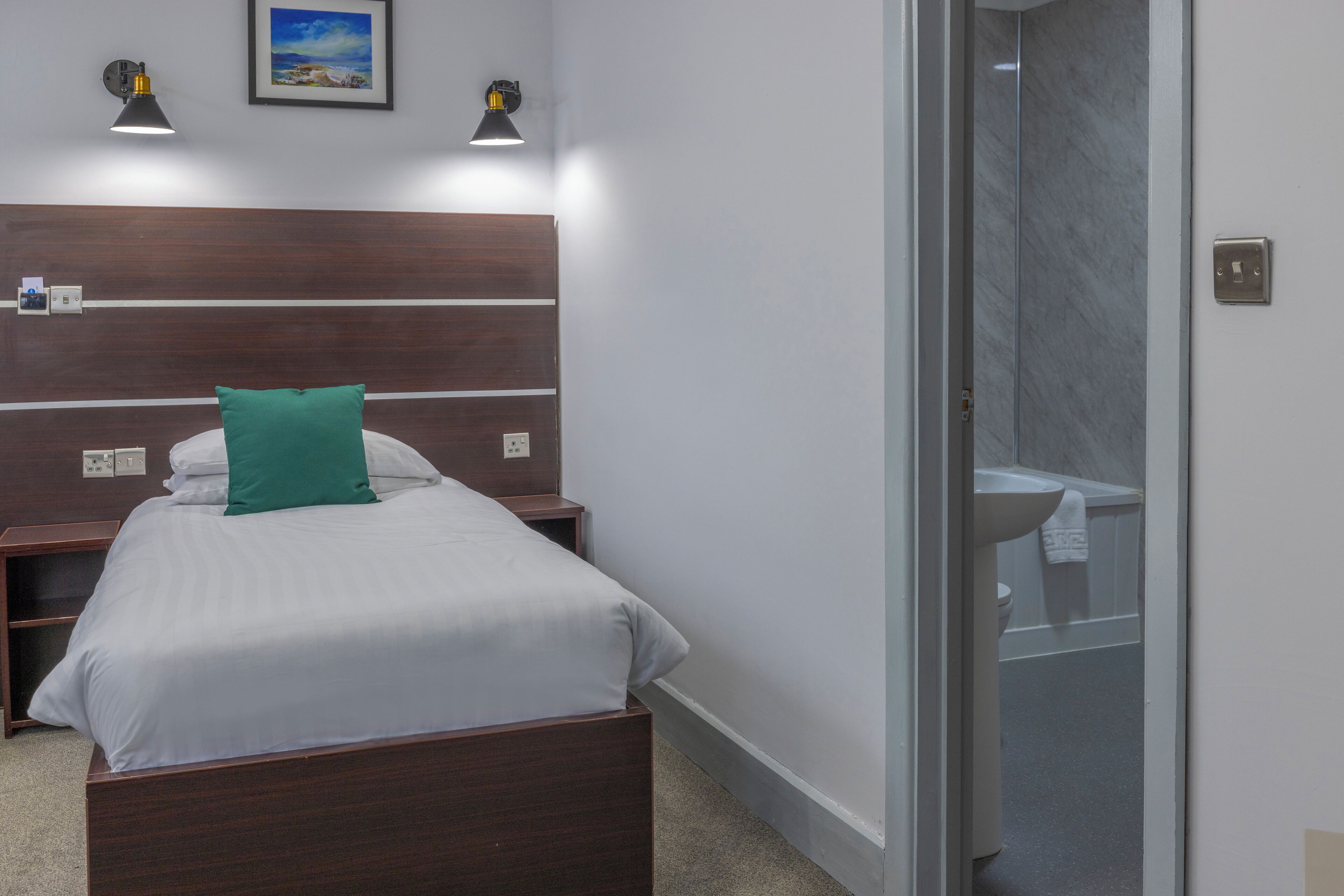 A hotel room with a wooden headboard, a bed with white bedding and a green pillow, two wall-mounted reading lamps, and a bathroom visible through a glass door.