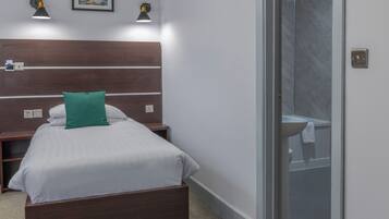 A hotel room with a wooden headboard, a bed with white bedding and a green pillow, two wall-mounted reading lamps, and a bathroom visible through a glass door.
