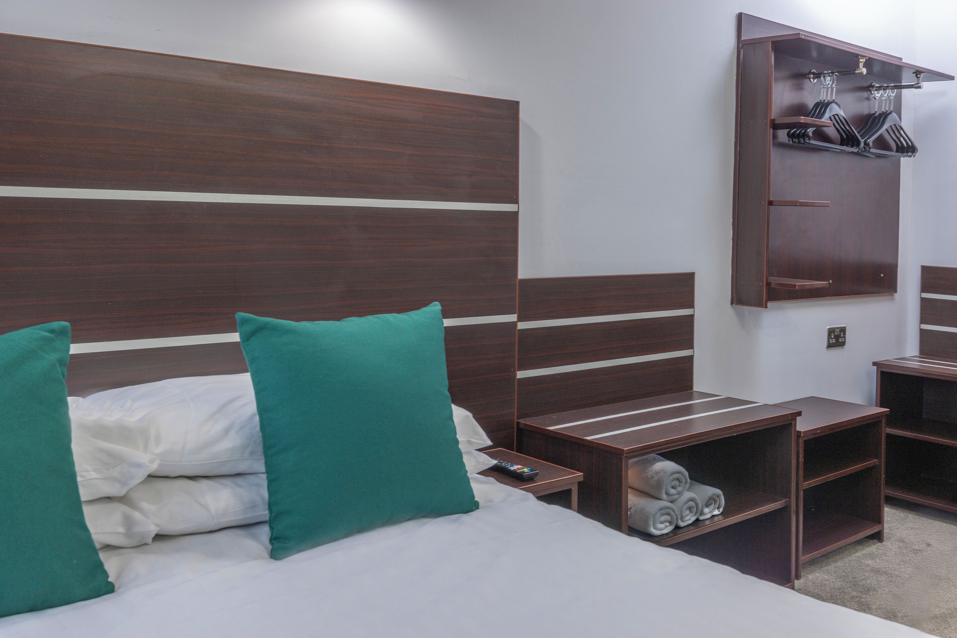 Habitación de hotel con una cama grande, dos almohadas verdes, un cabecero de madera, una mesita de noche con control remoto y un armario empotrado con espacio para colgar.