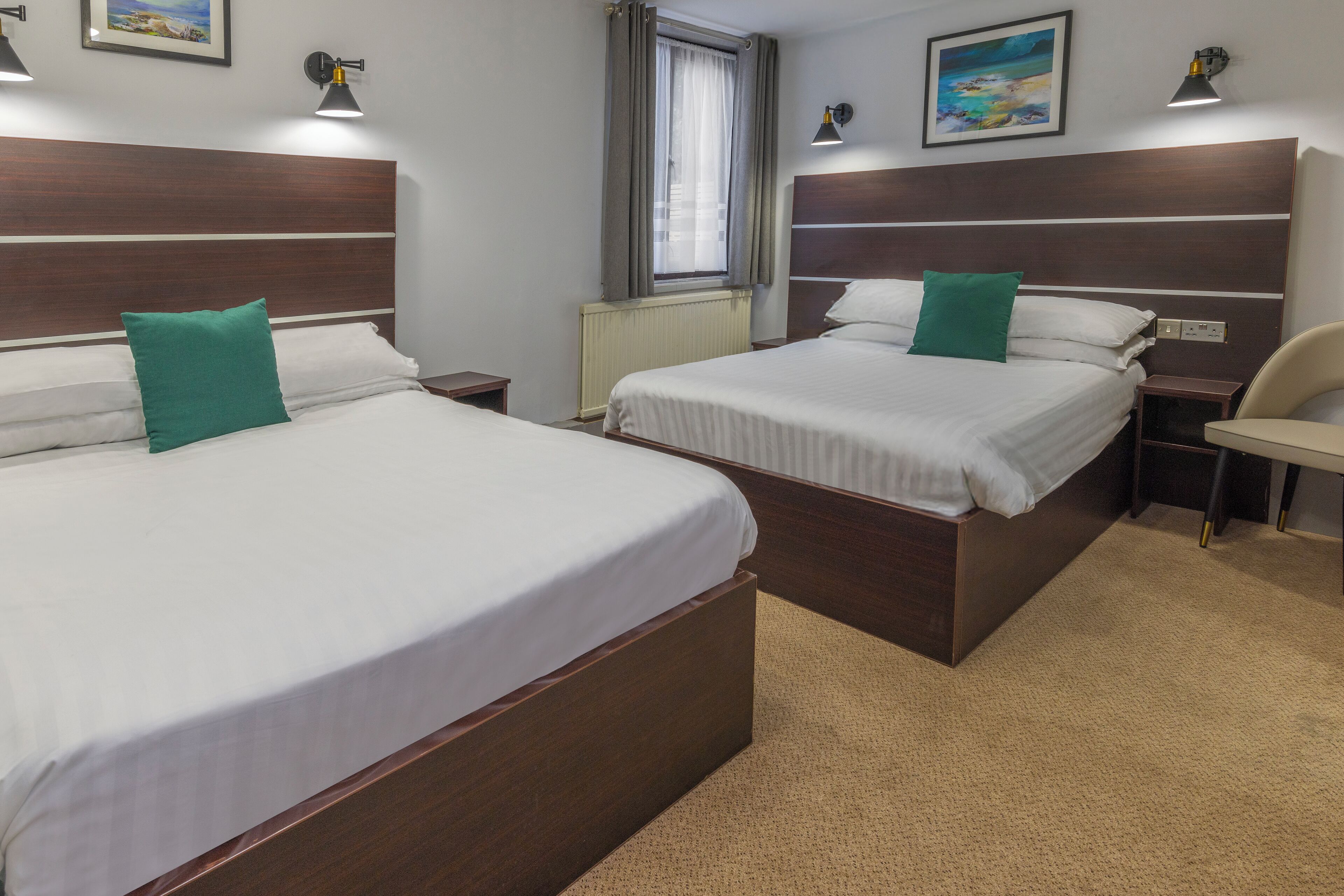 Una habitación de hotel con dos camas, una ventana con cortinas, un radiador y obras de arte en las paredes.