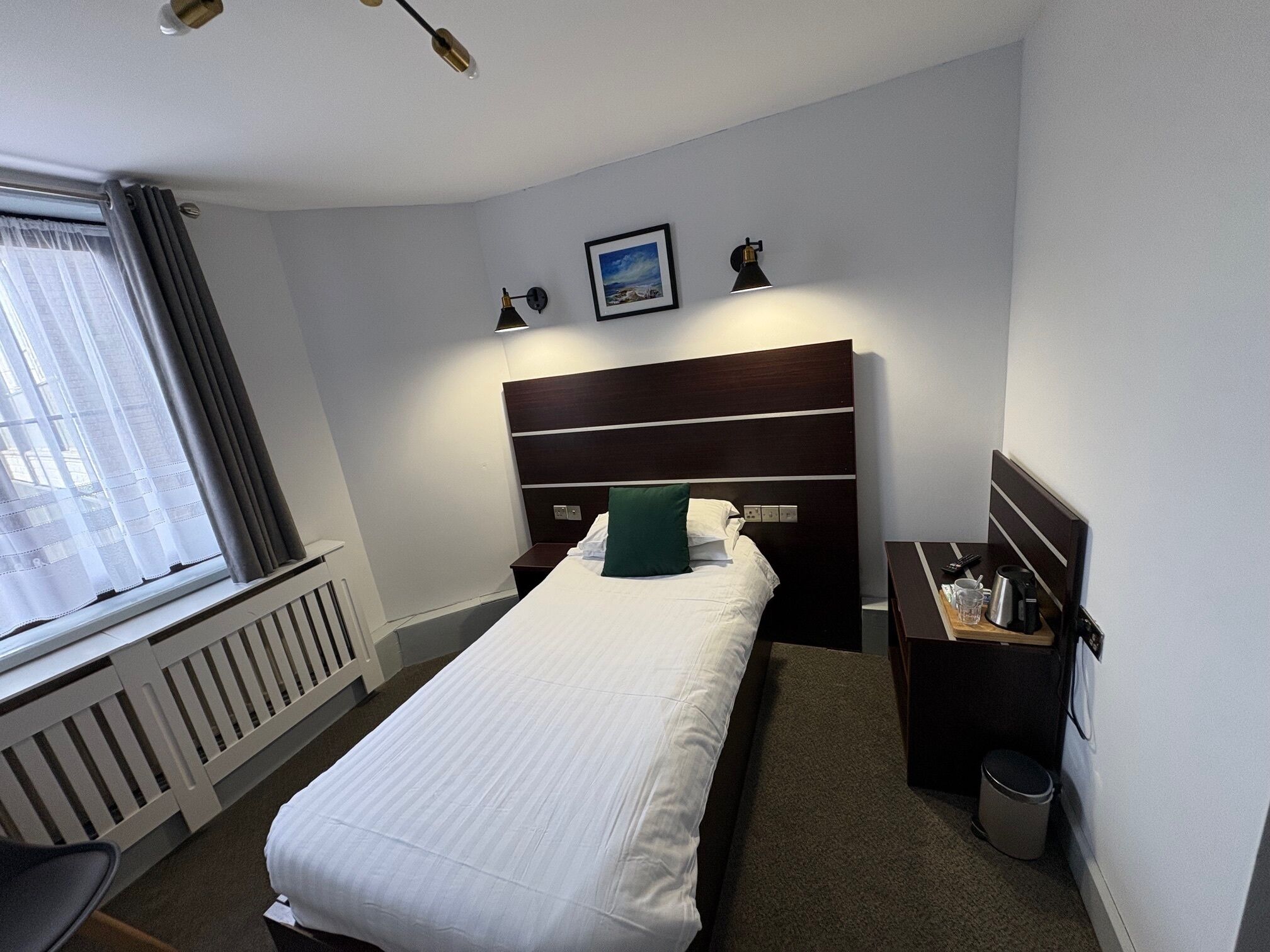 Une chambre d’hôtel bien rangée, avec un lit simple, une table de chevet avec une lampe, un petit tableau au mur et un radiateur.