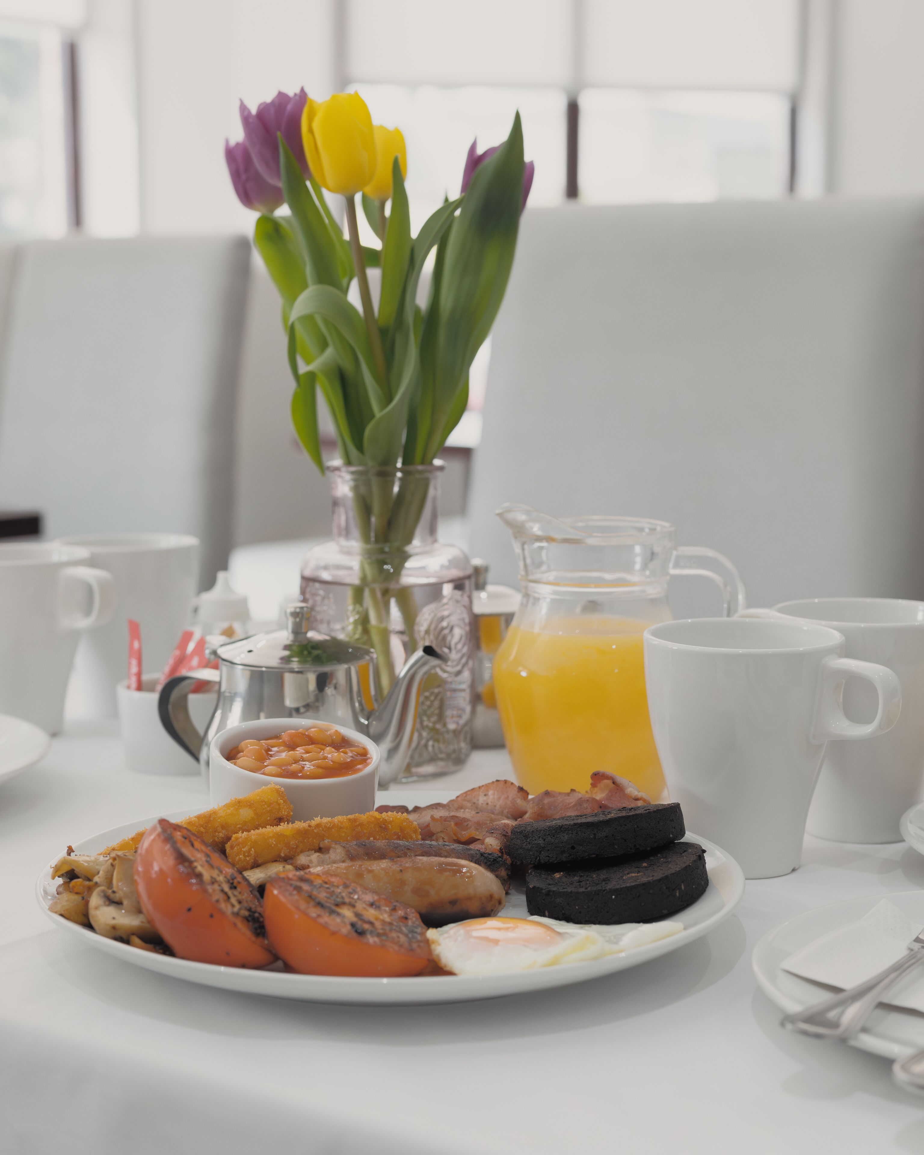 Une table de petit-déjeuner avec une assiette de nourriture, un pichet de jus d’orange et un vase de tulipes.
