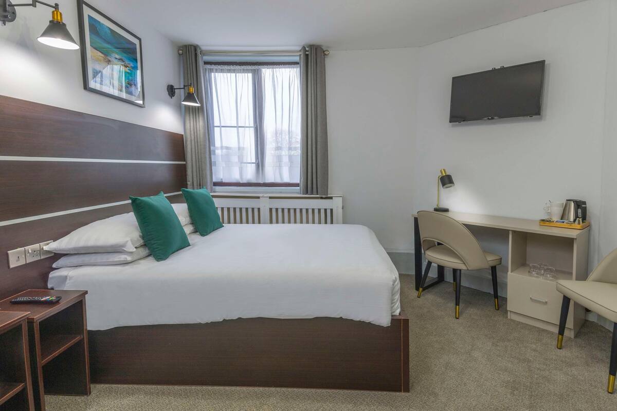 A hotel room with a bed, desk, chairs, a TV, and a window with curtains.