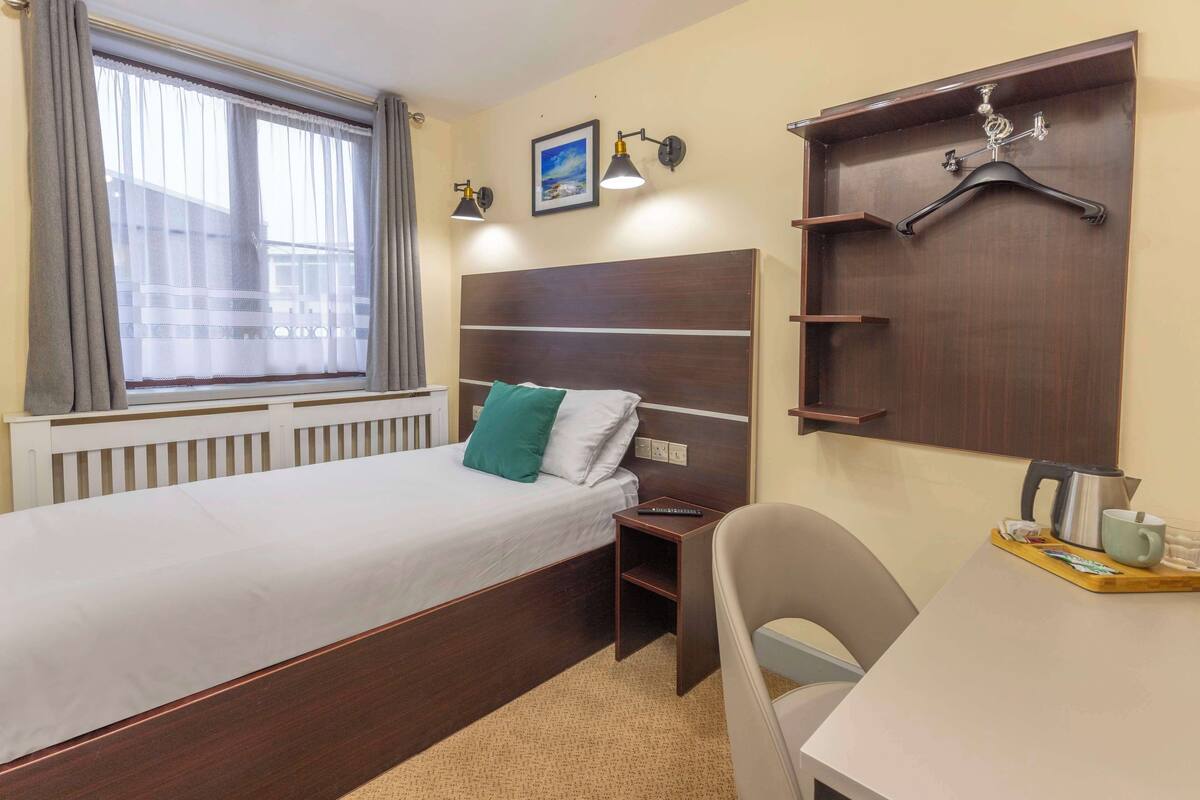 A neatly arranged hotel room with a single bed, bedside table, chair, and a small desk area with a kettle and mug.