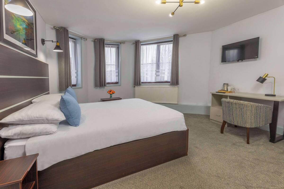A bedroom with a large bed, a desk, a chair, a TV, and two windows with curtains.