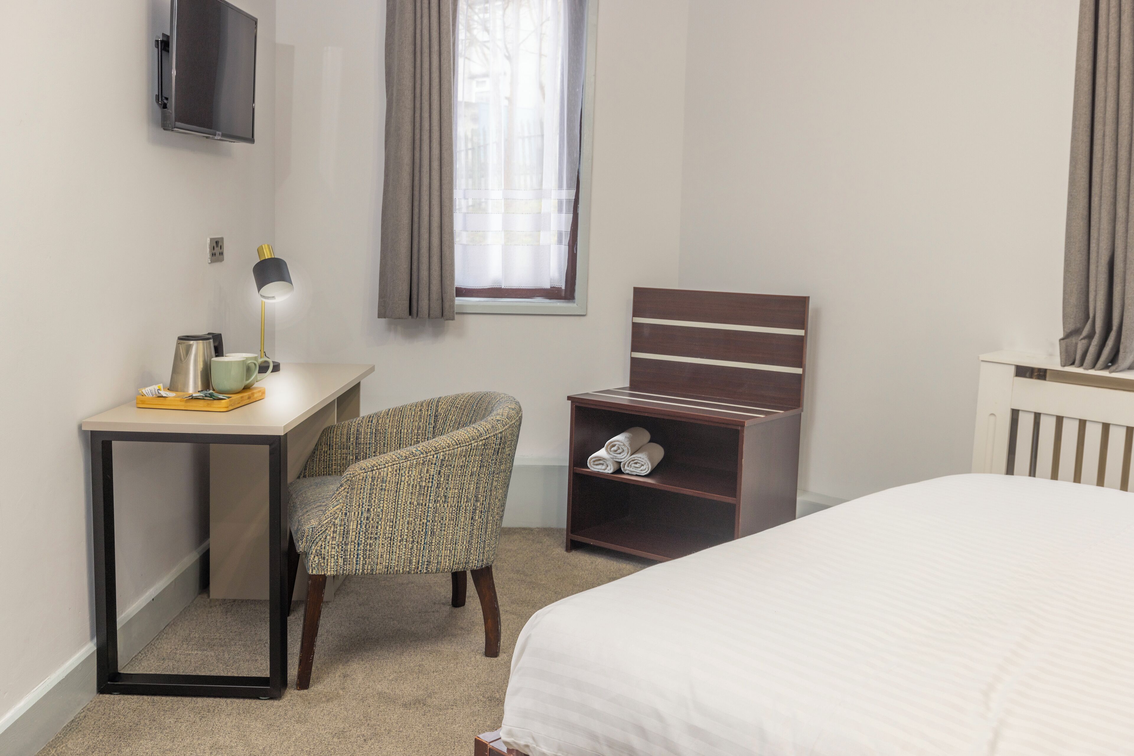 A hotel room with a bed, a desk with a lamp, a chair, a TV mounted on the wall, and a window with curtains.
