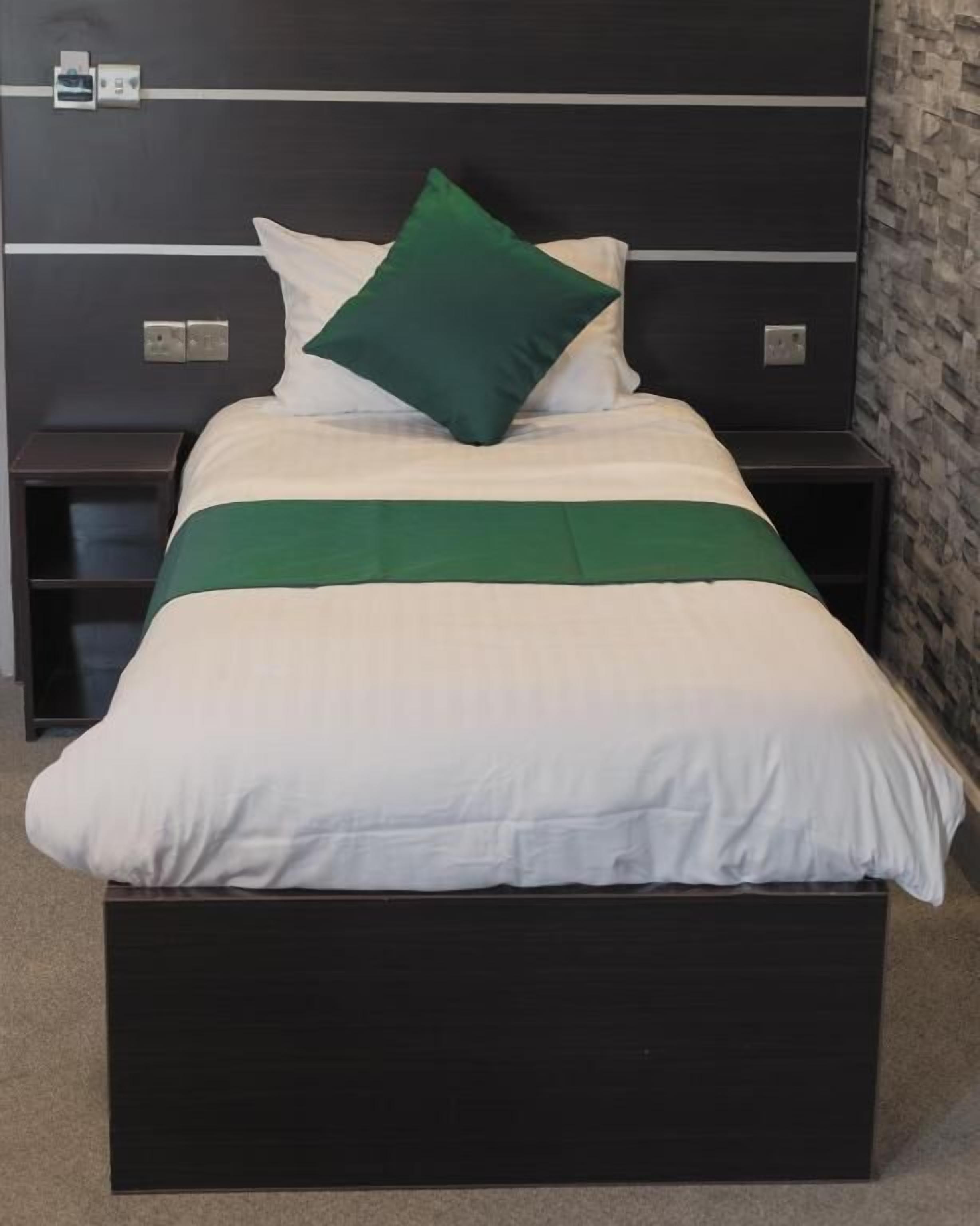A bed with a white and green bedspread, two white pillows, and a green accent pillow. There are two nightstands with a dark finish, each with a power outlet. The room has a dark wall with a textured pattern and a stone-like wall on the right.