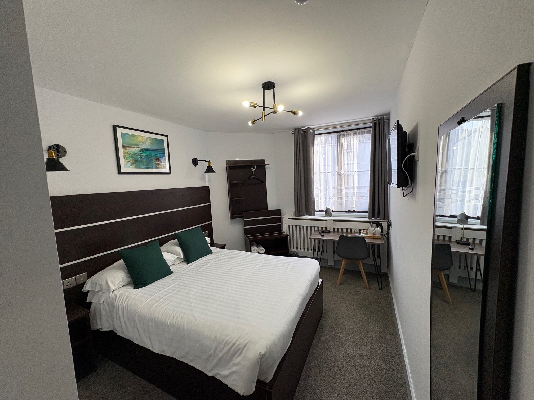 Une chambre d’hôtel avec un lit, un bureau, une chaise, un radiateur, une fenêtre avec des rideaux, un tableau au mur et un miroir.