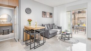 Un salon moderne avec un canapé gris, une table à manger en bois avec deux chaises, une petite table d’appoint avec un vase et un panier de fruits, et une grande fenêtre donnant sur un immeuble.