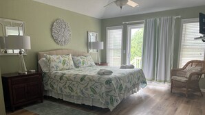 Ein Schlafzimmer mit einem Bett, Nachttischen, einer Kommode, einem großen Fenster mit Jalousien, einem Korbstuhl und einem Deckenventilator.