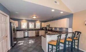 A modern kitchen with white cabinets, a central island with bar stools, and a stainless steel refrigerator. - Heart of Duck!!  Heated Pool, Hot Tub, Golf Cart  (Duck)