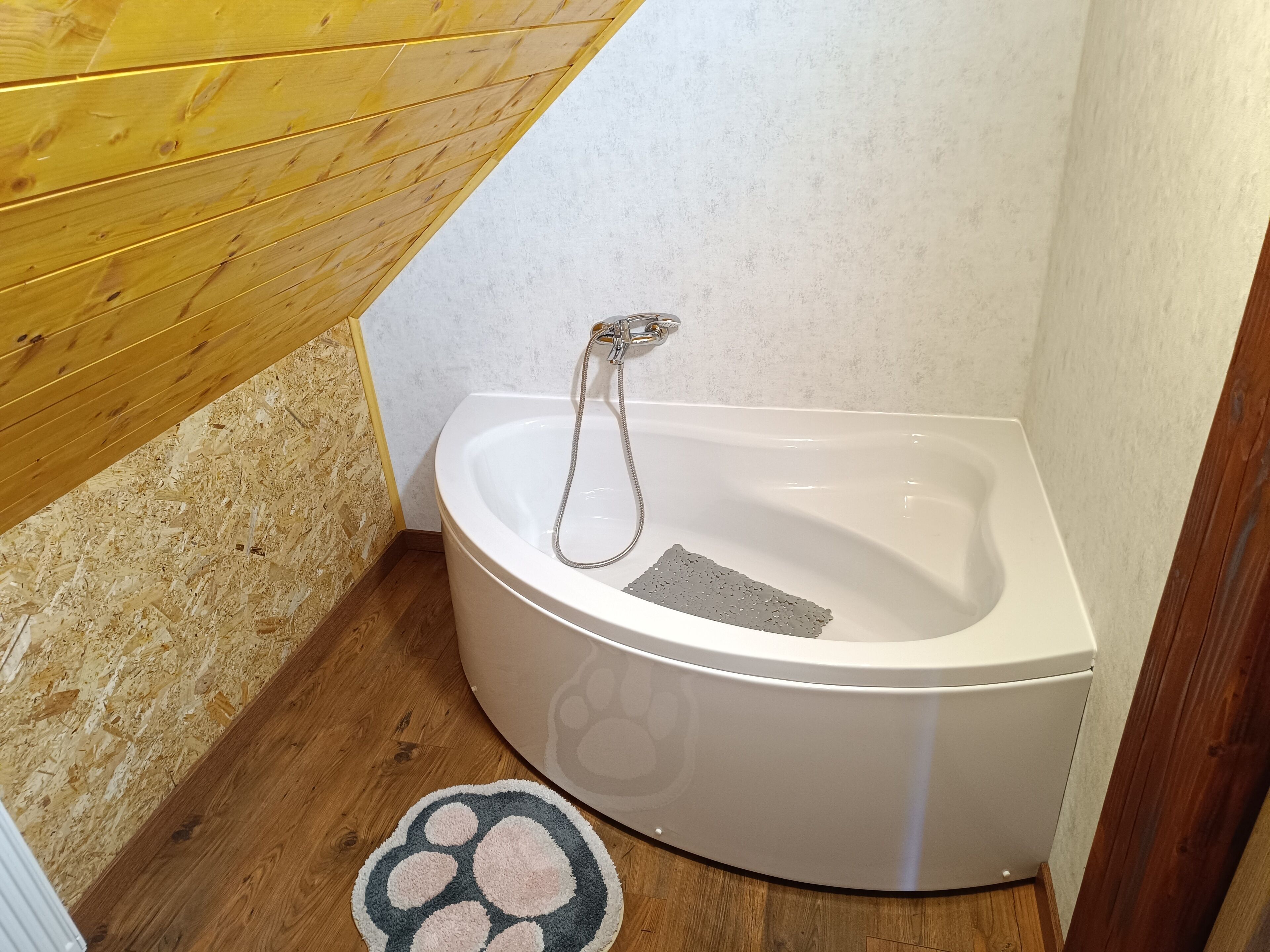 A corner bathroom with a white bathtub, wooden floor, and a pet paw print rug.