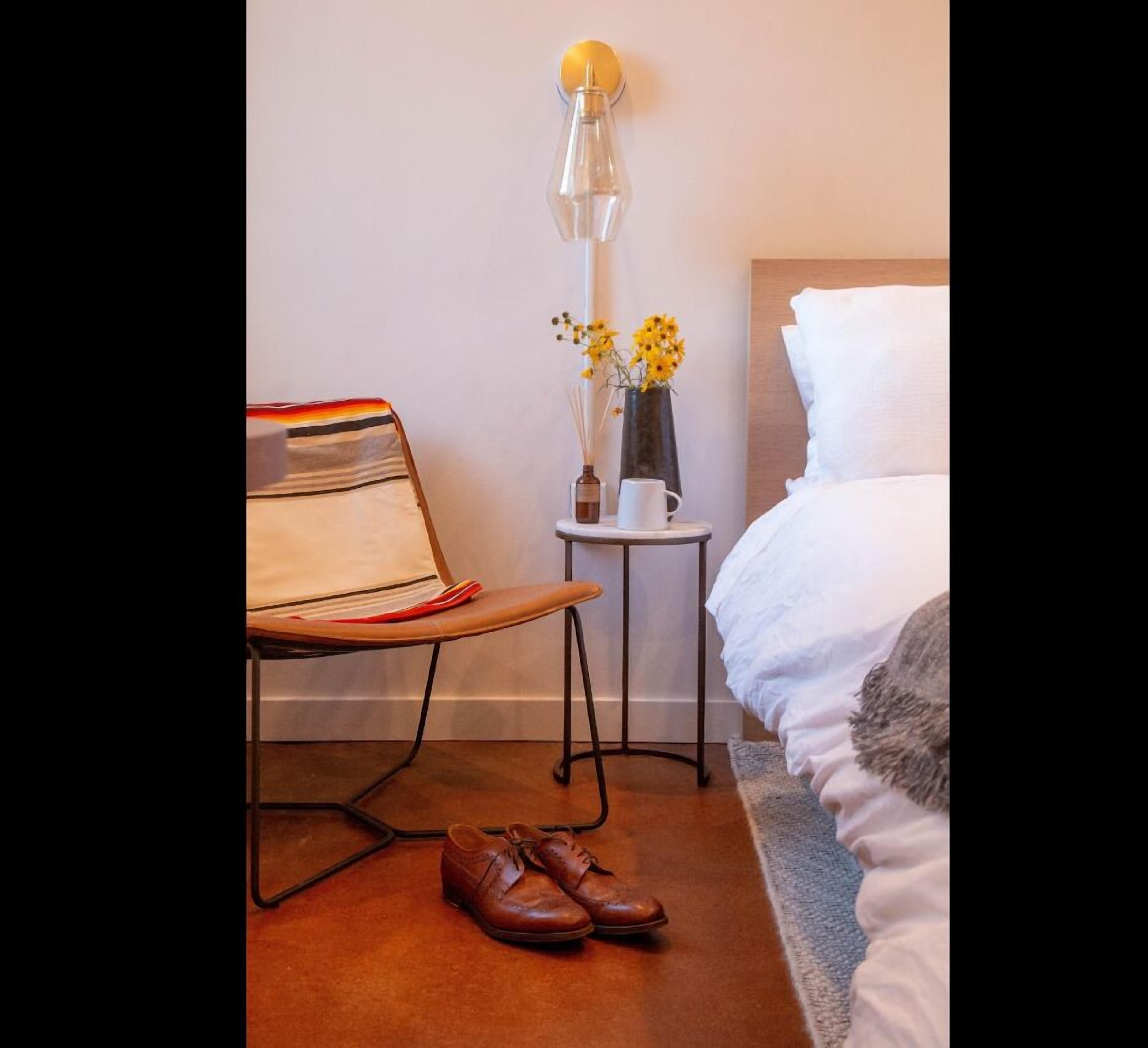 Habitación de hotel impecable con cama, mesita de noche con un jarrón de flores, lámpara y una silla con un libro.