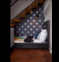 Um quarto de sótão pequeno e acolhedor, com teto inclinado, parede estampada, uma cama de solteiro com almofadas e uma escrivaninha com abajur.