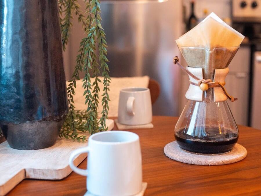 A coffee maker on a wooden table with a white mug and a potted plant.