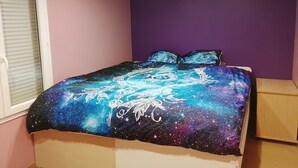 Ein Schlafzimmer mit einem Bett, das eine Bettdecke mit Galaxienthema hat, einem Fenster mit Jalousien und einer violetten Wand.