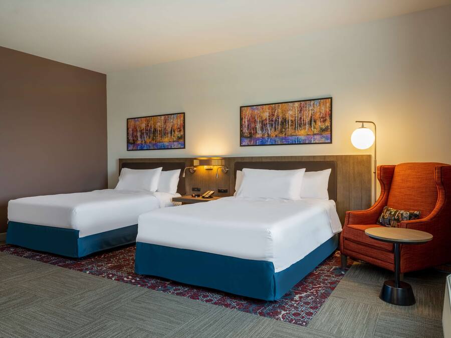 A hotel room with two beds, a chair, a small table, and two paintings on the wall.