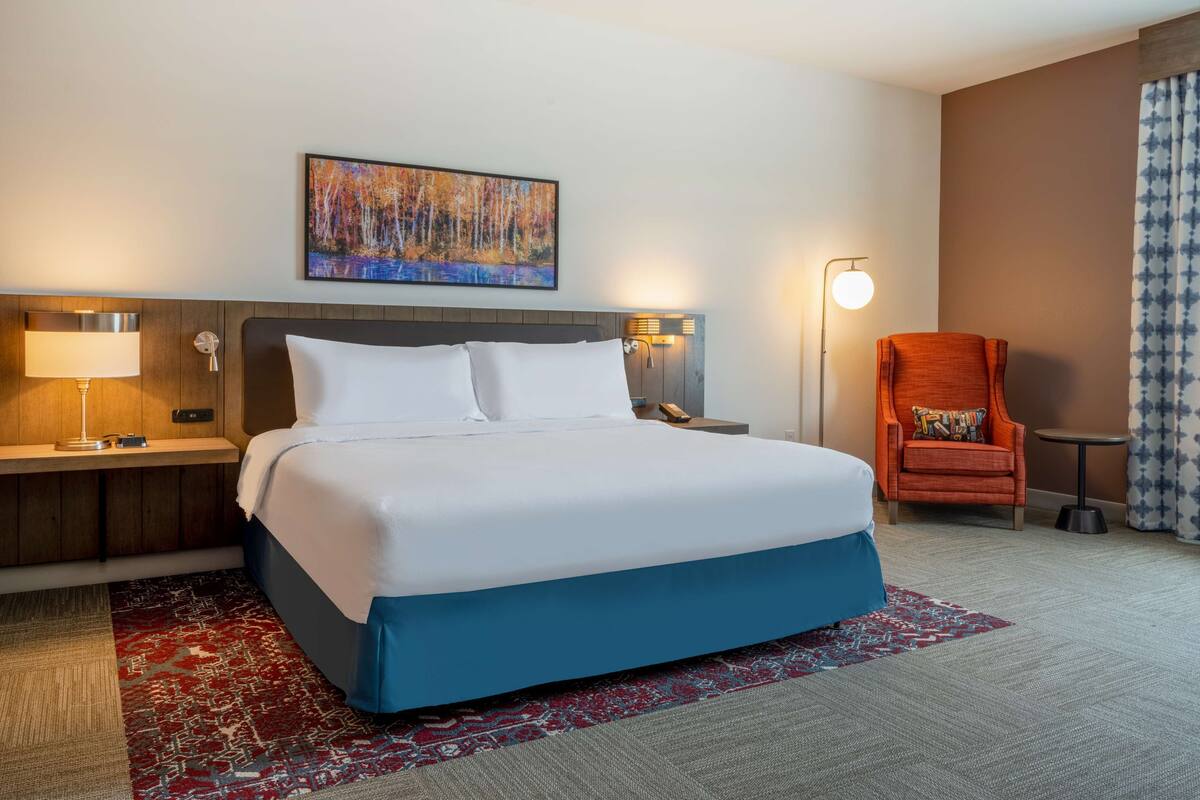 Een hotelkamer met een bed, nachtkastje, lamp, stoel en een schilderij aan de muur.