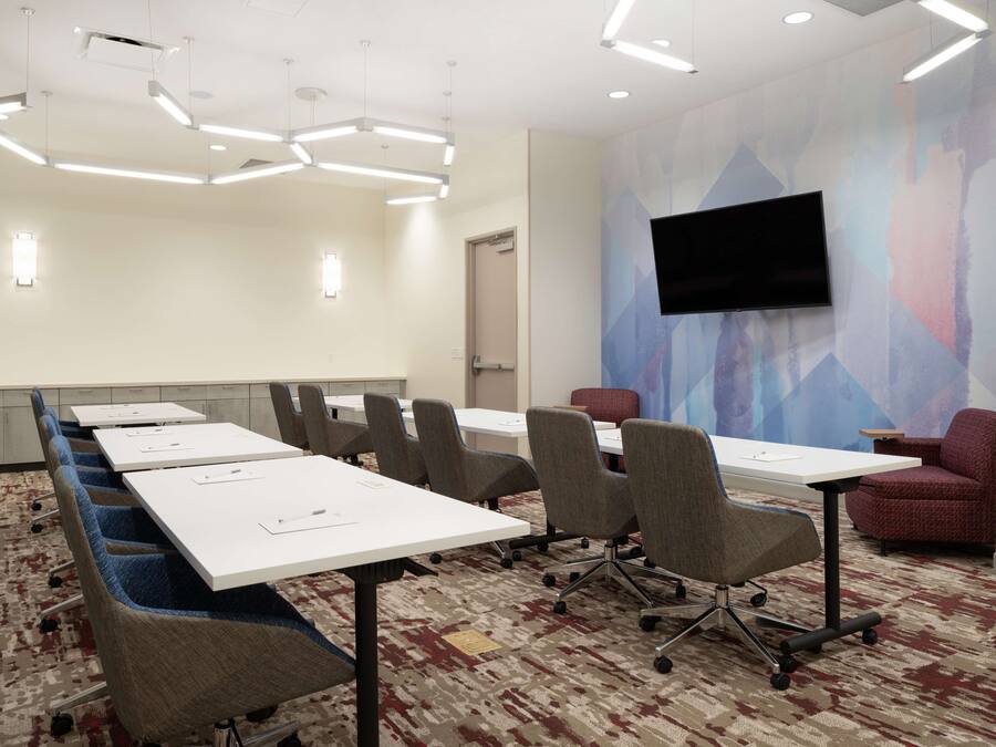 A conference room with white tables, grey chairs, a patterned carpet, a mounted TV, and a geometric wall mural.