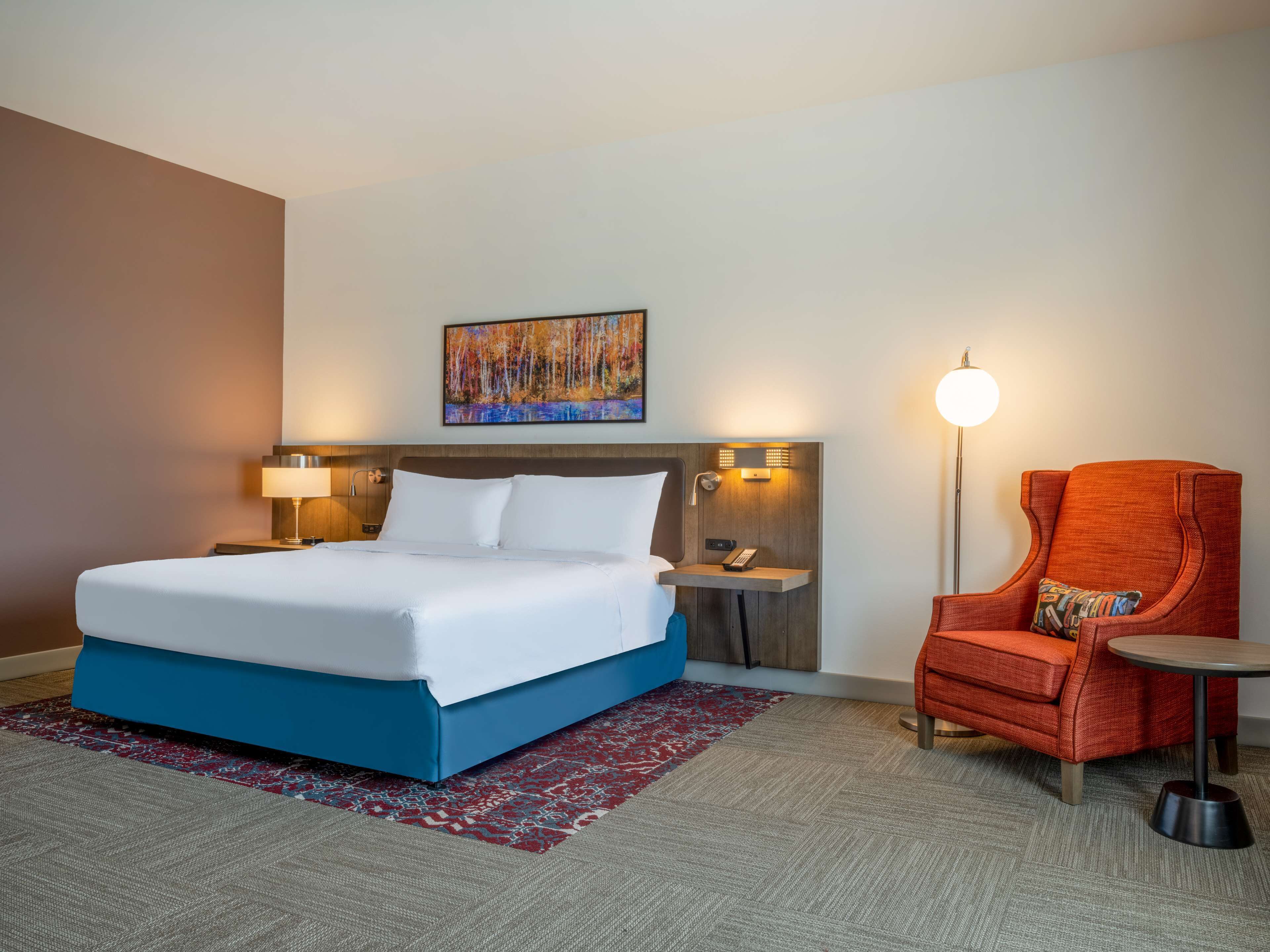 Una habitación de hotel con cama, una silla, una mesita, una lámpara y un cuadro en la pared.