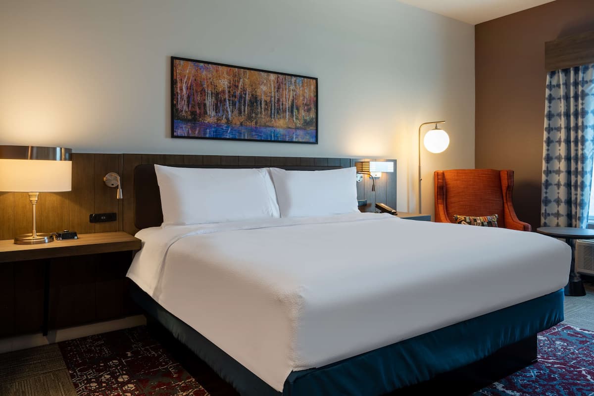 Een hotelkamer met een groot bed, een nachtkastje met lamp, een stoel, een schilderij aan de muur en een raam met gordijnen.