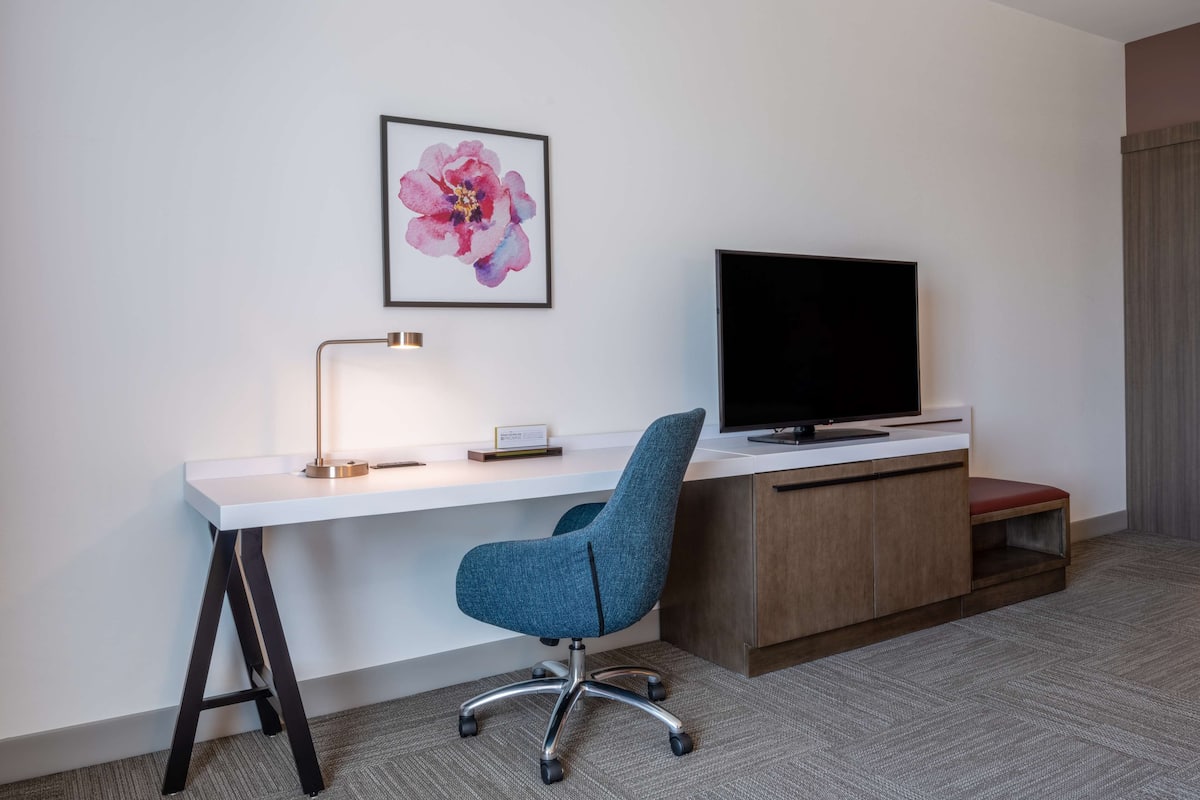 Hotelkamer met een wit bureau, een blauwe bureaustoel, een flatscreen televisie, een lamp en een bloemig kunstwerk in een lijst aan de muur.