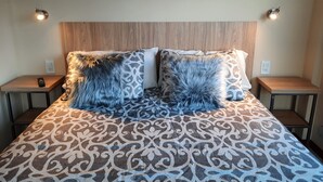 A bedroom with a patterned bedspread, two fluffy pillows, a wooden headboard, and a wall-mounted painting of flowers.