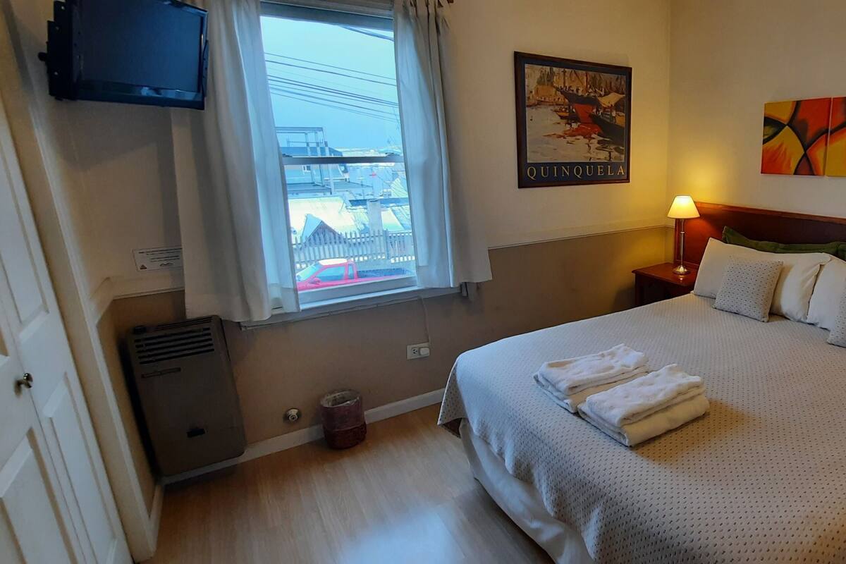 Ein Hotelzimmer mit einem Bett, zwei Nachttischen mit Lampen, einem Fenster mit Vorhängen, einem Wand-Fernseher, einem Gemälde und einem Heizkörper.