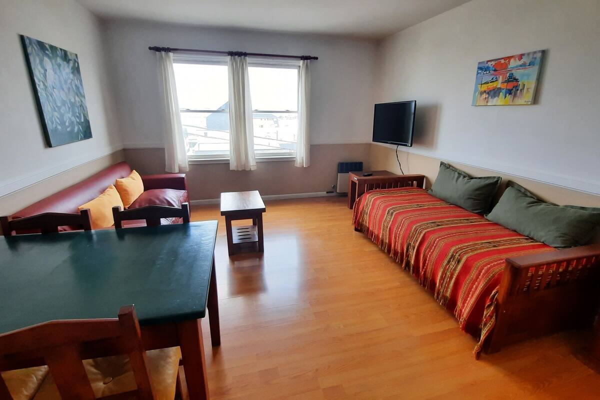 Ein Wohnzimmer mit einer Couch, einem Esstisch und Stühlen. Es gibt ein Fenster mit Vorhängen, einen an der Wand montierten Flachbildfernseher und ein Gemälde.