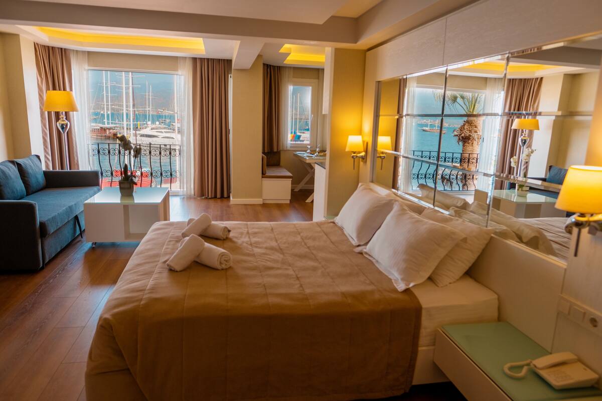 Une chambre d’hôtel avec un grand lit, un canapé, un bureau et un balcon donnant sur le port de plaisance.