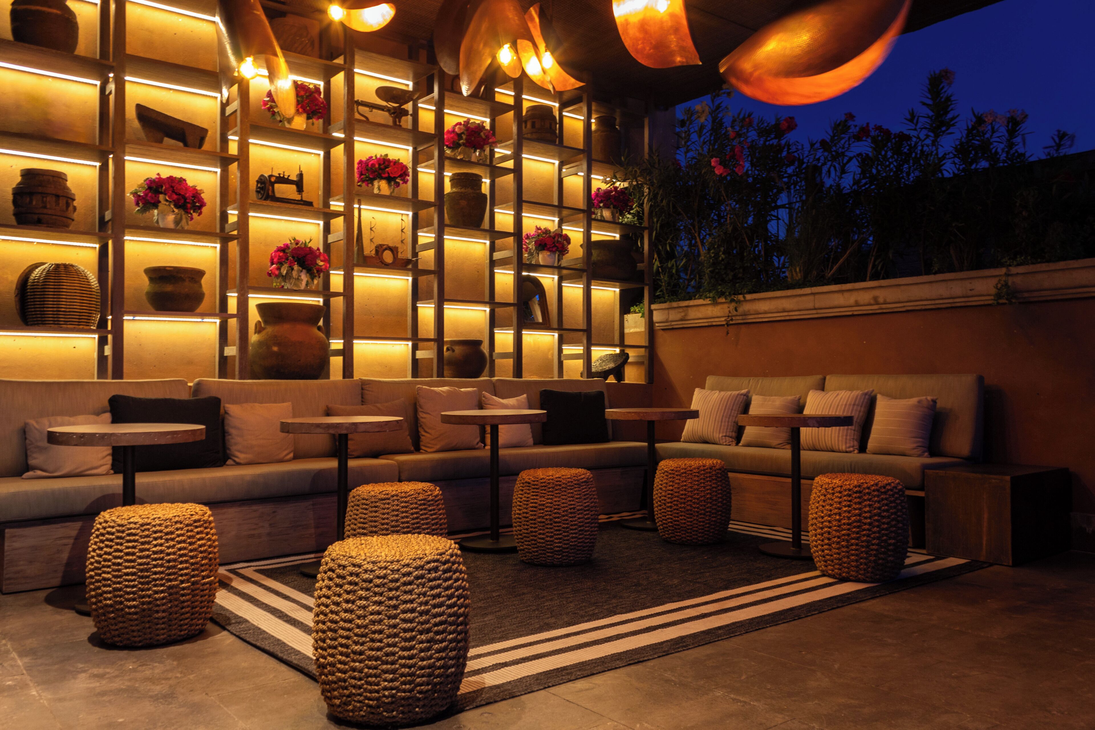 Un espacio lounge acogedor con sofás, mesas y otomanas de mimbre, iluminado con luz cálida y estantes con objetos decorativos.