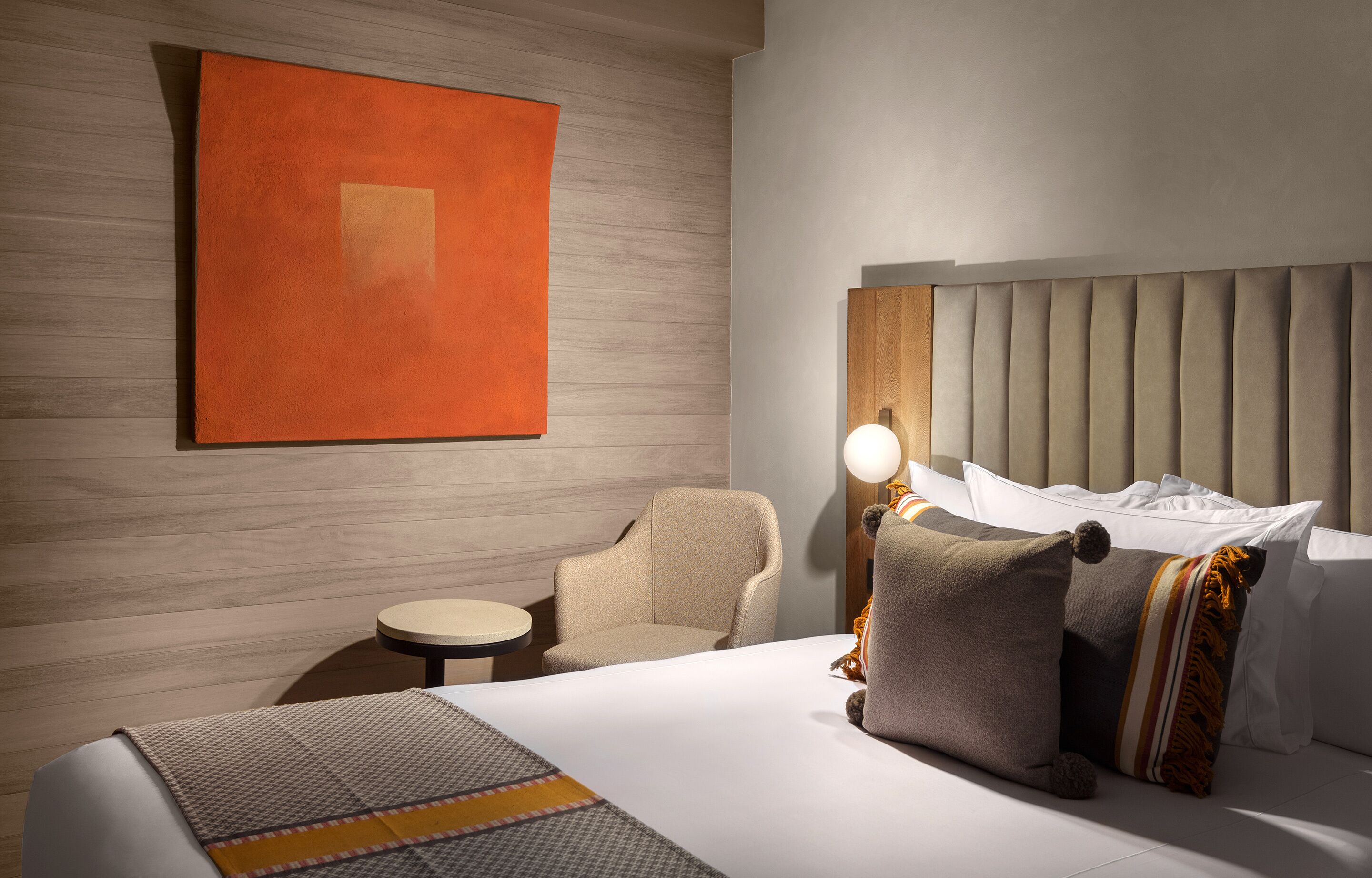 Habitación de hotel con una cama, una silla, una mesita, una lámpara y un cuadro grande en la pared.