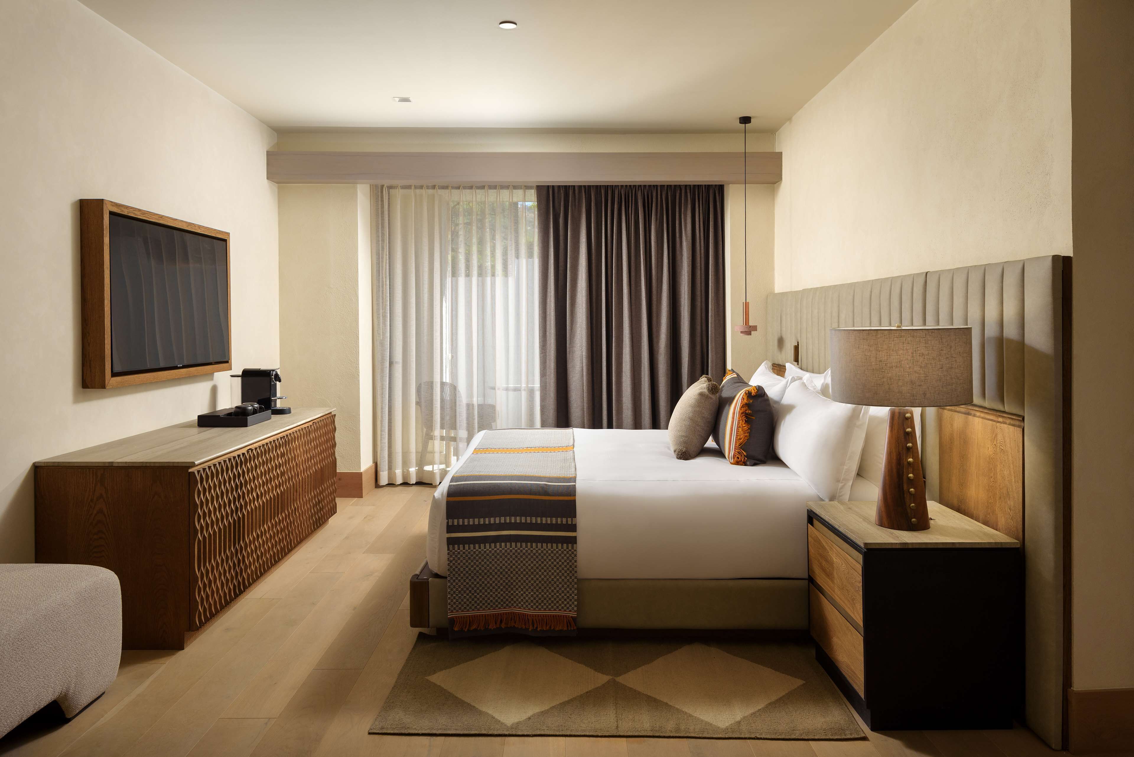 A modern hotel room with a large bed, a wooden dresser, a flat-screen TV, and a bedside table with a lamp.