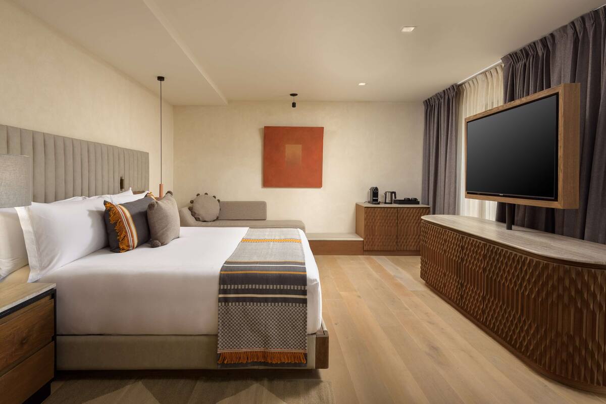 A modern hotel room with a large bed, a flat-screen TV, a wooden side table, and a small seating area with a sofa.